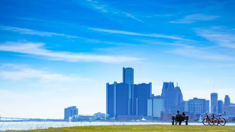 A couple sits on a bench looking over the river at the Detroit skyline